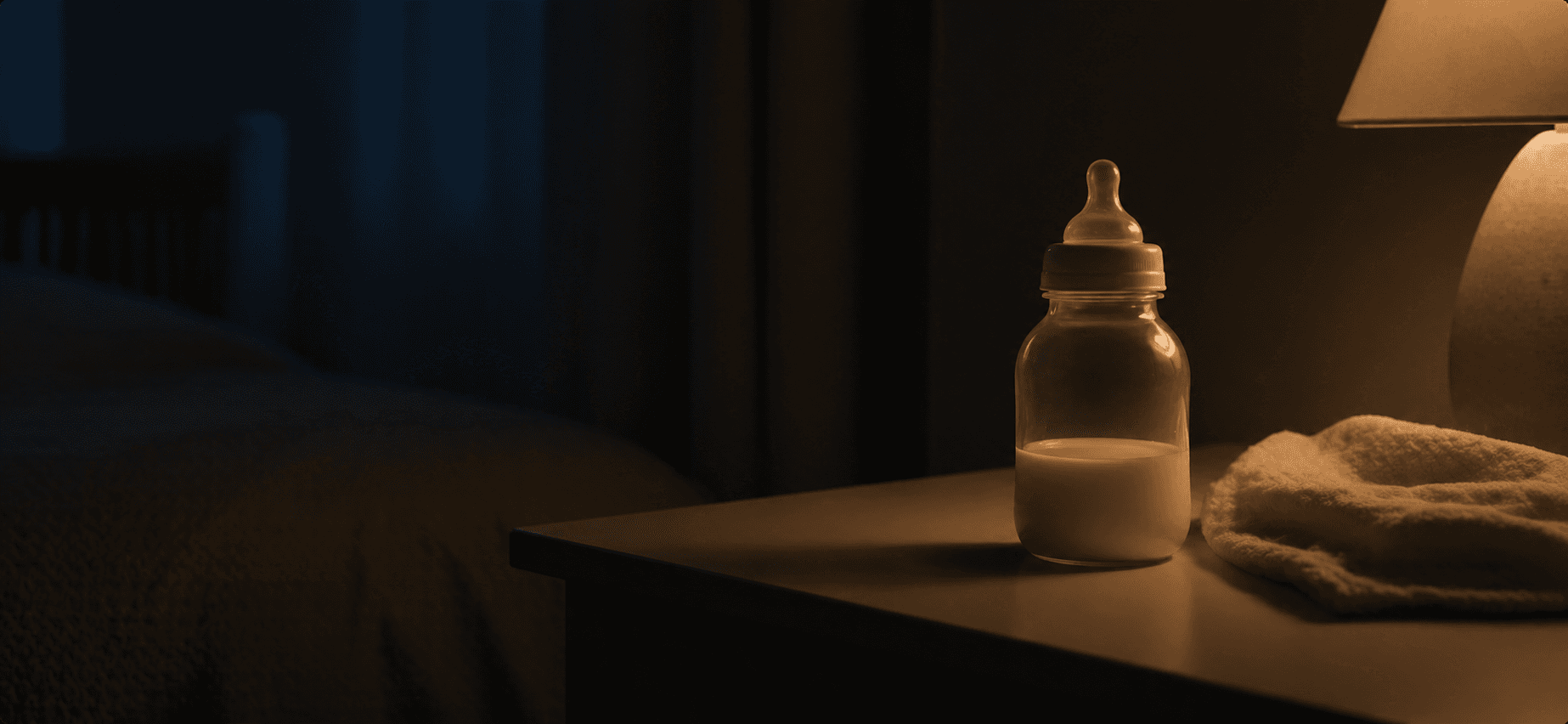 A quiet night-feed still life with a baby bottle on a bedside table under warm light.
